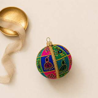 A round decorative ornament with colorful embroidered patterns in pink, blue, green, and yellow, featuring a central gold metallic band and a gold hanging loop. A small gold dish and a beige ribbon are placed beside it on a light cream background.
