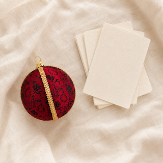 A red and black damask-patterned Christmas ornament with a gold braided trim and hanging loop, styled on a beige linen fabric next to a stack of blank cream-colored cards.