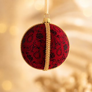 A round red Christmas ornament with a black damask pattern and a gold braided center stripe, hanging against a warm golden bokeh background.