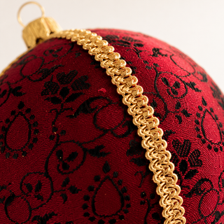 A close-up of a red Christmas ornament with a black floral damask pattern and a shiny gold braided trim running diagonally across it.