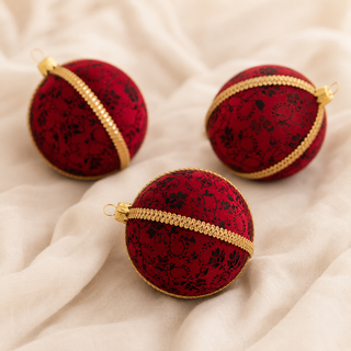 Three red Christmas ornaments with a black floral pattern and gold trim, arranged on a draped cream fabric background.