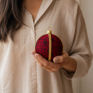 A person in a beige linen shirt holds a red and black patterned spherical ornament with a gold hanging loop and gold vertical trim.