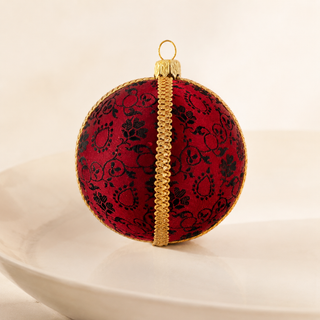 A spherical red Christmas ornament with a black damask pattern and a vertical gold trim, resting on a white ceramic plate against a warm beige background.