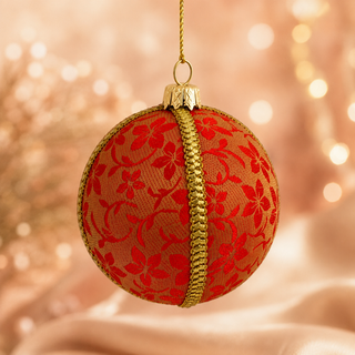 A round red and gold Christmas ornament hanging from a gold string, featuring a red floral pattern on a gold textured background with a vertical gold braided stripe down the center, set against a warm, softly blurred bokeh background.