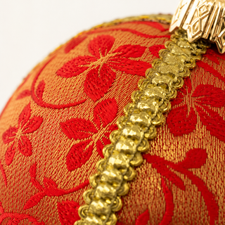 Close-up of a decorative spherical ornament with a red floral pattern woven onto a warm orange-gold fabric, accented by a shiny gold braided trim and a gold hanging loop.