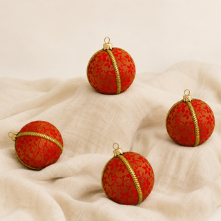Four red patterned Christmas ornaments with gold trim and gold hanging loops, arranged on draped cream fabric against a light neutral background.