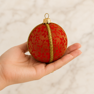 A hand holding a round red and gold Christmas ornament with a floral pattern and a gold center stripe, against a light marble background.