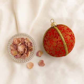 A decorative spherical ornament in red and gold patterned fabric with a gold braided band and gold hanging loop, styled on a white fabric background next to a small glass dish of dried rose petals.