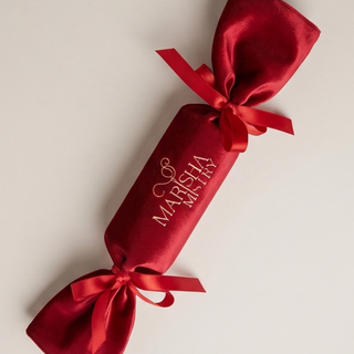 A red satin Christmas cracker ornament with gold foil text reading 'MARRY MARISHA' and tied with matching red satin ribbons at both ends, shown on a light neutral background.