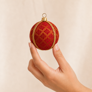 A hand holding a round red Christmas ornament with a gold patterned design and gold trim against a soft beige background.