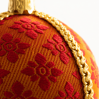 Close-up of a decorative spherical ornament featuring a red floral pattern woven onto a warm orange-gold fabric surface, with a gold braided trim and a gold hanging cap visible at the top.