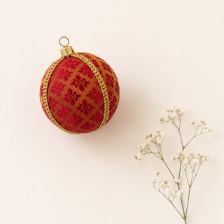 A round red and gold embroidered Christmas ornament with a gold hanging loop, styled next to small white baby's breath flowers on a warm off-white background.