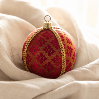 A round red and gold embroidered Christmas ornament with a gold braided trim and a metal hanging loop, resting on draped cream fabric.