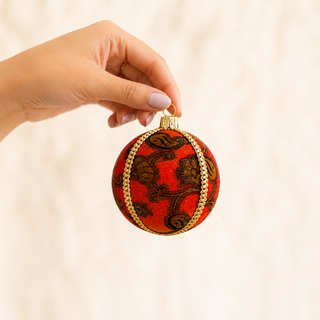 A hand with light pink manicured nails holds a round red Christmas ornament featuring a brown paisley pattern and gold trim, set against a soft, warm beige background.
