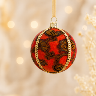 A round red Christmas ornament with a gold chain pattern and dark brown scrollwork, hanging from a gold ribbon against a warm, blurred bokeh background.