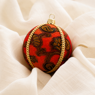 A round red Christmas ornament with a gold and brown paisley pattern and gold braided trim, resting on draped cream fabric.