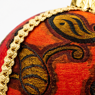 Close-up of a decorative spherical ornament featuring a rich red and orange embroidered pattern with black detailing and a braided gold trim.