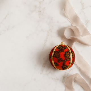A round red Christmas ornament with a brown floral pattern and gold trim, resting on a white marble surface next to a cream ribbon.