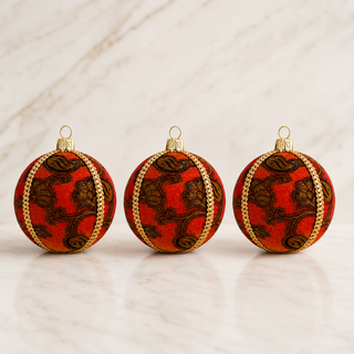 Three red Christmas baubles with gold braided trim and a dark brown floral scroll pattern, arranged on a white marble surface.