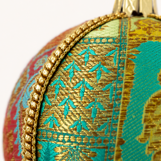 Close-up of a decorative spherical ornament featuring intricate gold and turquoise woven patterns, a braided gold trim, and a gold hanging loop against a plain white background.