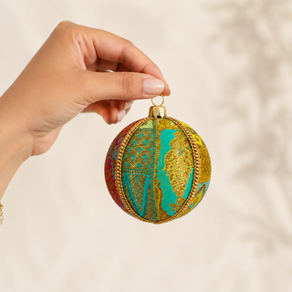 A hand holding a spherical decorative ornament with a gold and turquoise map design, set against a soft beige background with subtle leaf shadows.