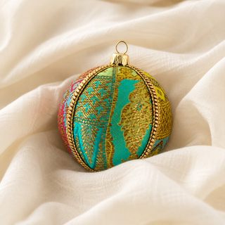 A decorative spherical ornament with a gold-tone hanging loop, featuring a turquoise and gold patterned center with multicolored side panels and a gold braided border, resting on draped cream fabric.