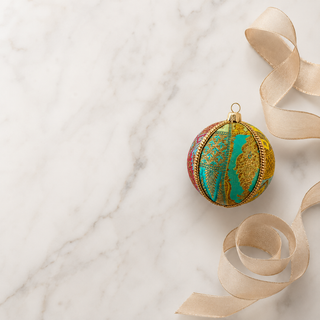 A decorative Christmas ornament shaped like a globe, featuring a gold and turquoise map design with red accents, styled on a white marble surface with a beige ribbon.
