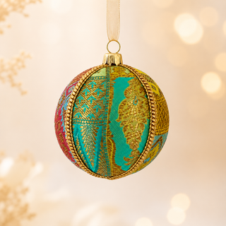 A spherical Christmas ornament hanging from a cream ribbon, decorated with gold metallic patterns and borders, turquoise blue panels, and pink accents, set against a warm, softly blurred bokeh background.