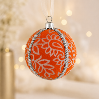 An orange Christmas ornament with a white floral pattern and silver trim, hanging by a white ribbon against a warm, softly blurred bokeh background.