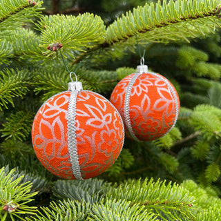 Two orange and silver fabric baubles hanging on a green fir tree branch in an outdoor setting