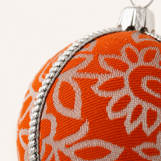 Close-up of an orange Christmas ornament with a white floral pattern and a silver metallic trim and hanger.