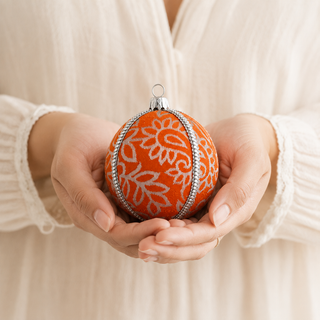 A person in a cream-colored ribbed top holds a decorative orange Christmas ornament with a white floral pattern and a silver metallic trim.
