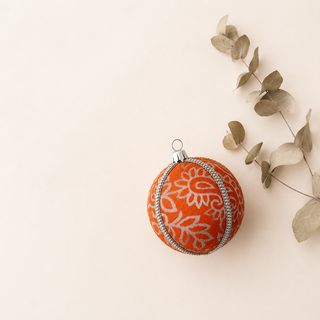 An orange Christmas ornament with a white floral pattern and silver trim, styled on a light beige background with dried eucalyptus branches.