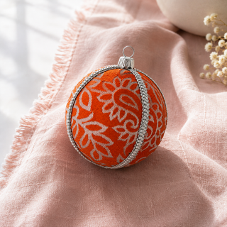 An orange Christmas ornament with a white floral pattern and silver trim, resting on a soft pink textured fabric.