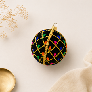 A decorative black Christmas ornament with a gold wire grid pattern and colorful embroidered dots, styled on a light beige background with dried flowers and a gold bowl.