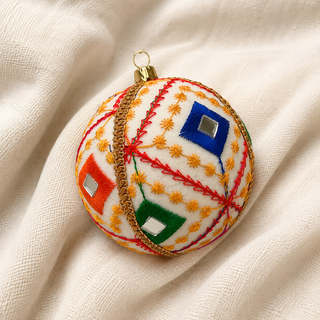 Sohan fabric bauble styled on linen, a 10cm handmade ornament with bold red diagonal lines with a fine gold star-stitch pattern and mirror-centred diamonds.