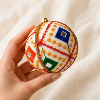Sohan bauble hanging with soft bokeh lights, showing its crisp white ground and bold red diagonal lines with a fine gold star-stitch pattern and mirror-centred diamonds.