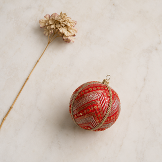 Subhash fabric bauble styled on linen, an 8cm handmade ornament with diagonal bands of gold and silver embroidery with scattered sequin florets and a beaded gold trim.