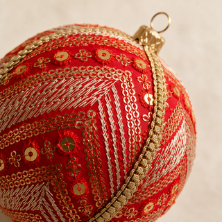 Subhash bauble hanging with soft bokeh lights, showing its deep red silk ground and diagonal bands of gold and silver embroidery with scattered sequin florets and a beaded gold trim.
