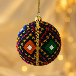 A spherical Christmas ornament hanging from a gold string, decorated with intricate multicolor embroidery on a dark navy base, featuring red, orange, green, and yellow geometric patterns against a warm, blurred golden bokeh background.