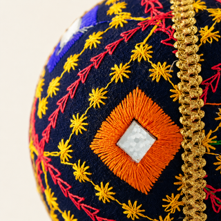 Close-up of a decorative embroidered oval textile piece, showing a navy base with yellow starbursts, red arrow motifs, an orange diamond patch with a white center, and a gold braided trim on a light background.