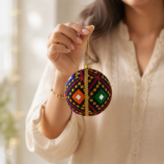 A person in a cream blouse holds up a round, hand-woven Christmas ornament. The ornament features a vibrant multicolor pattern of red, yellow, blue, green, and orange geometric designs with gold accents.