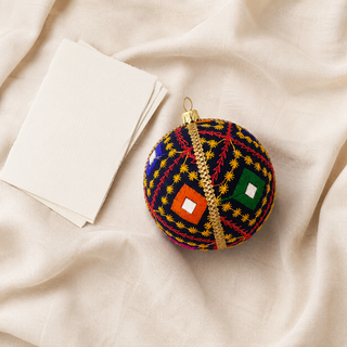 A round embroidered Christmas ornament with a gold hanging loop, featuring a navy blue base with red, yellow, and green geometric patterns, styled on a beige fabric surface next to blank cards.
