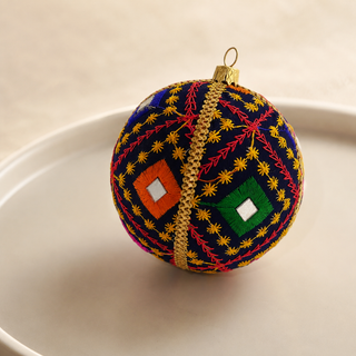 A spherical embroidered ornament with a navy blue base, gold floral patterns, red and green diamond motifs, and a gold hanging loop, displayed on a light beige surface.