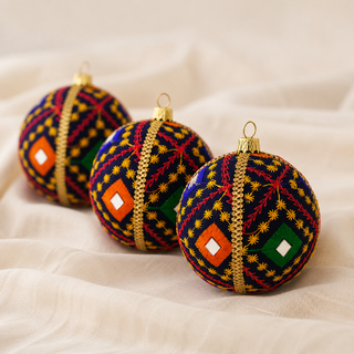 Three decorative Christmas ornaments shaped like spheres, featuring intricate multicolor embroidery in blue, red, yellow, green, and orange with gold metallic accents, displayed on draped cream fabric.