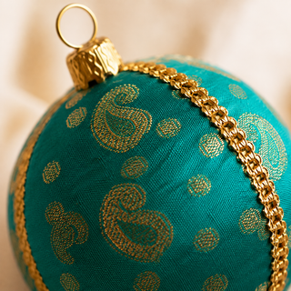 Tarla bauble hanging with soft bokeh lights, showing its deep teal ground and gold paisley motifs and delicate dotted brocade with a gold cord seam trim.