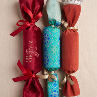 Three decorative Christmas crackers arranged vertically on a light beige fabric background. The left cracker is deep red satin with a red ribbon and gold 'MARIA' script.