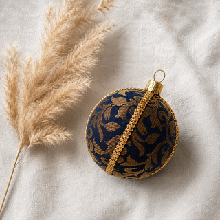 Vani fabric bauble styled on linen, a 10cm handmade ornament with sweeping gold floral scrollwork woven throughout with a gold cord seam trim.