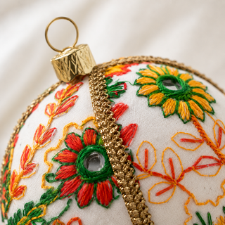 Close-up of the Vanika bauble showing embroidered floral medallions in orange, yellow, green and red with mirror-work centres and a gold cord trim on cream reclaimed fabric.