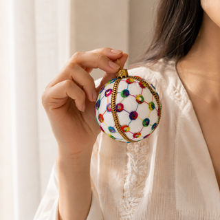 Vikram bauble hanging with soft bokeh lights, showing its cream ground and a lattice of coloured thread with rainbow stitched circles and tiny mirror centres.